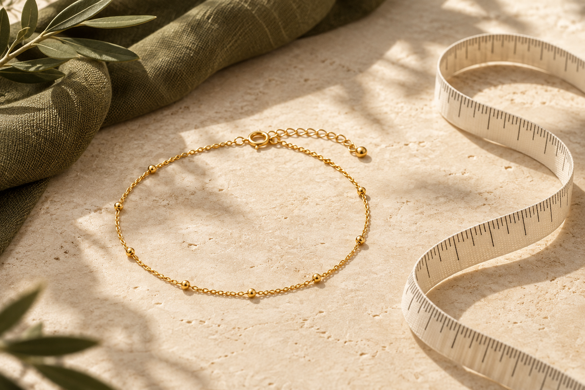 Illustrative gold-coloured anklet beside an unmarked measuring ribbon on cream stone and olive linen