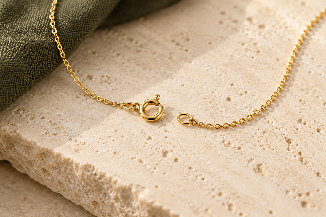 Illustrative gold-coloured chain with an open spring-ring clasp and end loop on cream stone beside olive linen