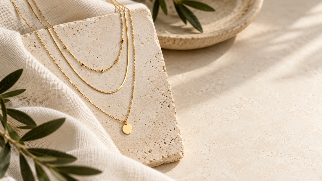 Illustrative gold-coloured necklaces at staggered lengths on a cream display beside olive leaves.
