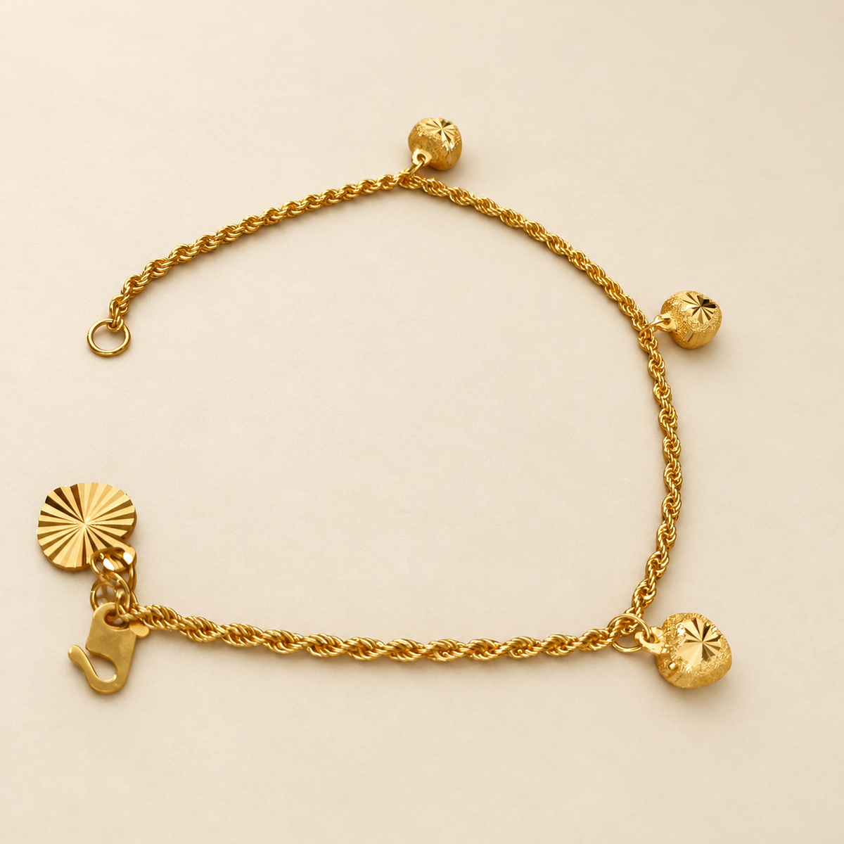 Full view of the 916 gold hollow rope bracelet with three textured charms, hook clasp and flat end tag on ivory